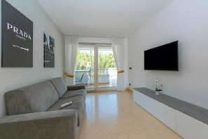 A modern living room with a grey sofa, a flat-screen TV mounted on the wall, and a sliding glass door leading to an outdoor area.