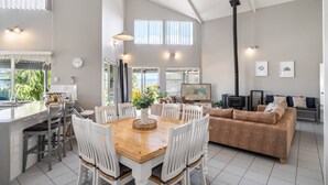A spacious living area with a wooden dining table, white chairs, a brown sofa, and a large window offering a view of greenery.