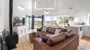 A spacious living area with a large brown leather sofa, a wooden coffee table, a black wood-burning stove, a flat-screen TV, and a kitchen area with white cabinets and a countertop.