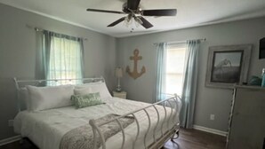 Un dormitorio con cama, ventilador de techo, mesita de noche, cómoda y ventana con cortinas.