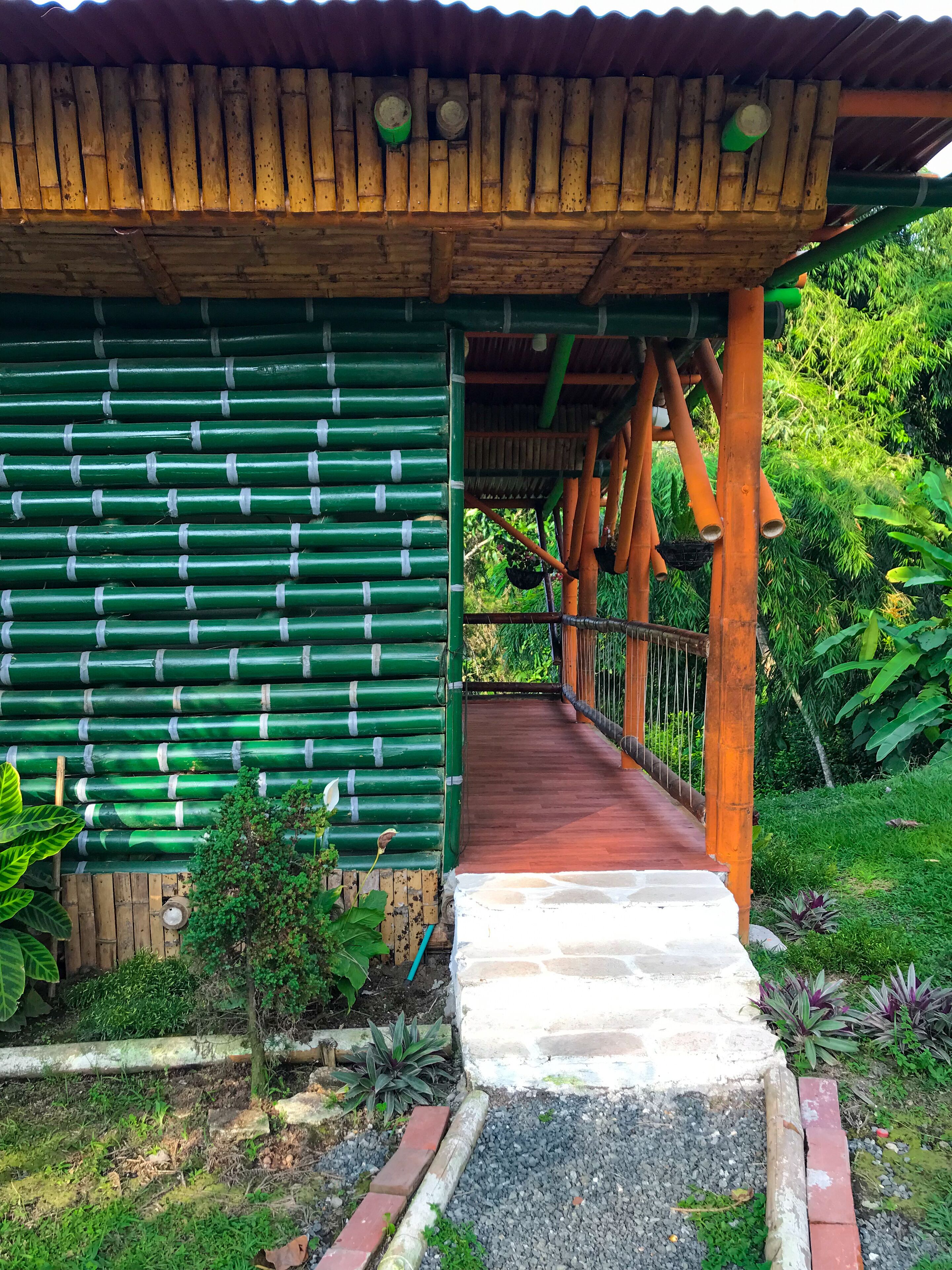 Une structure en bois couverte, dotée d’un mur en bambou vert, d’un chemin rouge menant à l’intérieur et d’un jardin luxuriant.