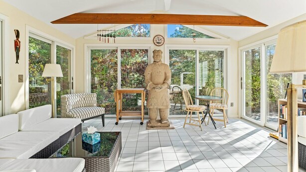 A spacious room with a high ceiling, large windows, a stone statue, a white sofa, a wicker coffee table, and a wooden dining table with chairs. - Bike trail, Tennis Court, Surrounded by nature (Eastham)
