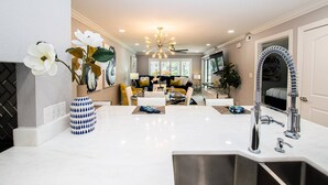 A modern kitchen with a white countertop, stainless steel sink, and a unique faucet. There is a dining area in the background with a sofa and a TV. - King Suite & Patio | Ground Floor | Monthly Stay | 1BR, Pool, Near Tampa | Saddlebrook Resort (Wesley Chapel)