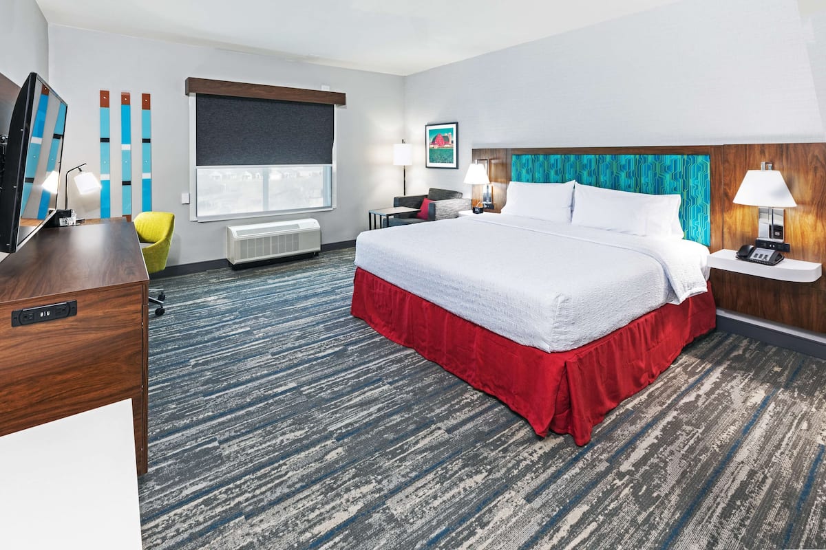 A hotel room with a large bed, a desk with a computer, a chair, a window with blinds, a wall-mounted TV, and a wall-mounted telephone.