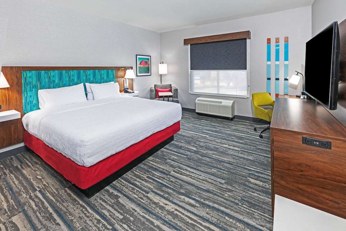 A hotel room with a large bed, a desk, a chair, a TV, and a window with blinds.