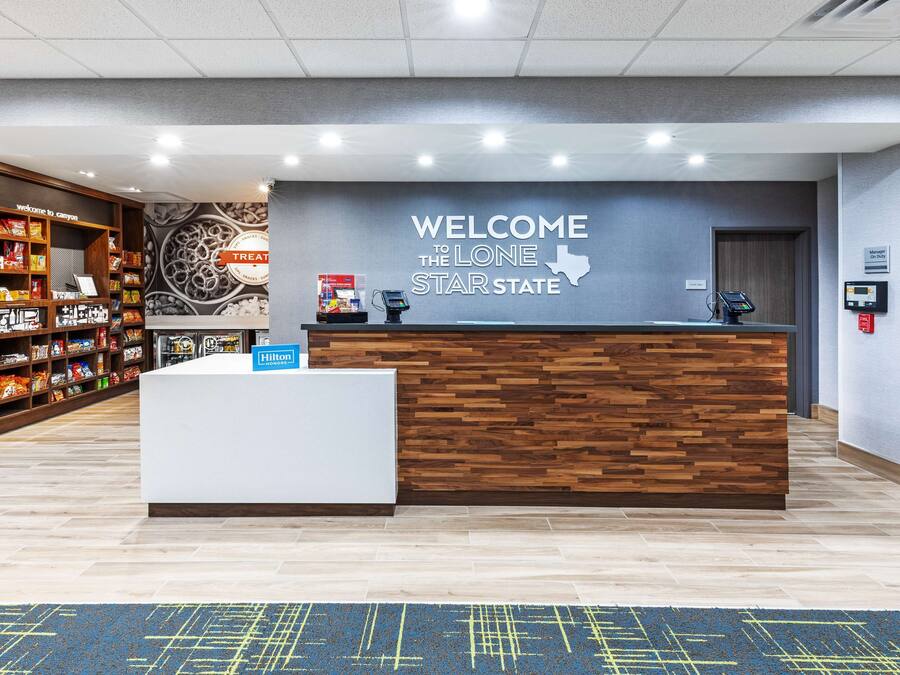 Un área de recepción de hotel con un mostrador de madera, un cartel en la pared que dice "WELCOME TO THE LONE STAR STATE" y un quiosco de autoservicio.