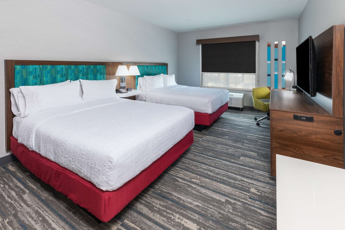 A hotel room with two beds, a desk, a TV, and a window with blinds.