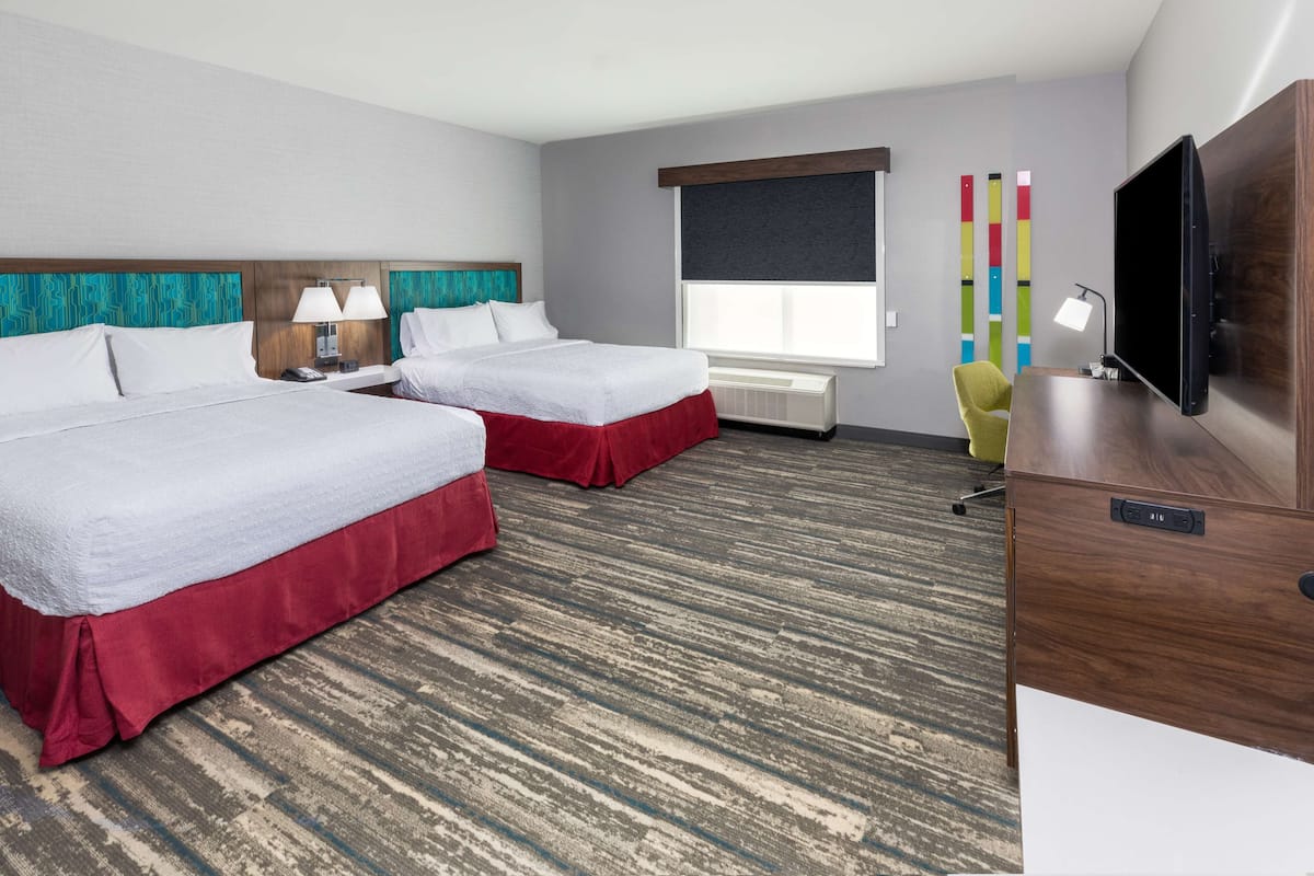 A hotel room with two beds, a desk, a TV, and a window with blinds.