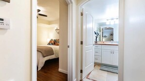 Corredor que dá para um quarto com cama e ventilador de teto, e um banheiro com pia e espelho.