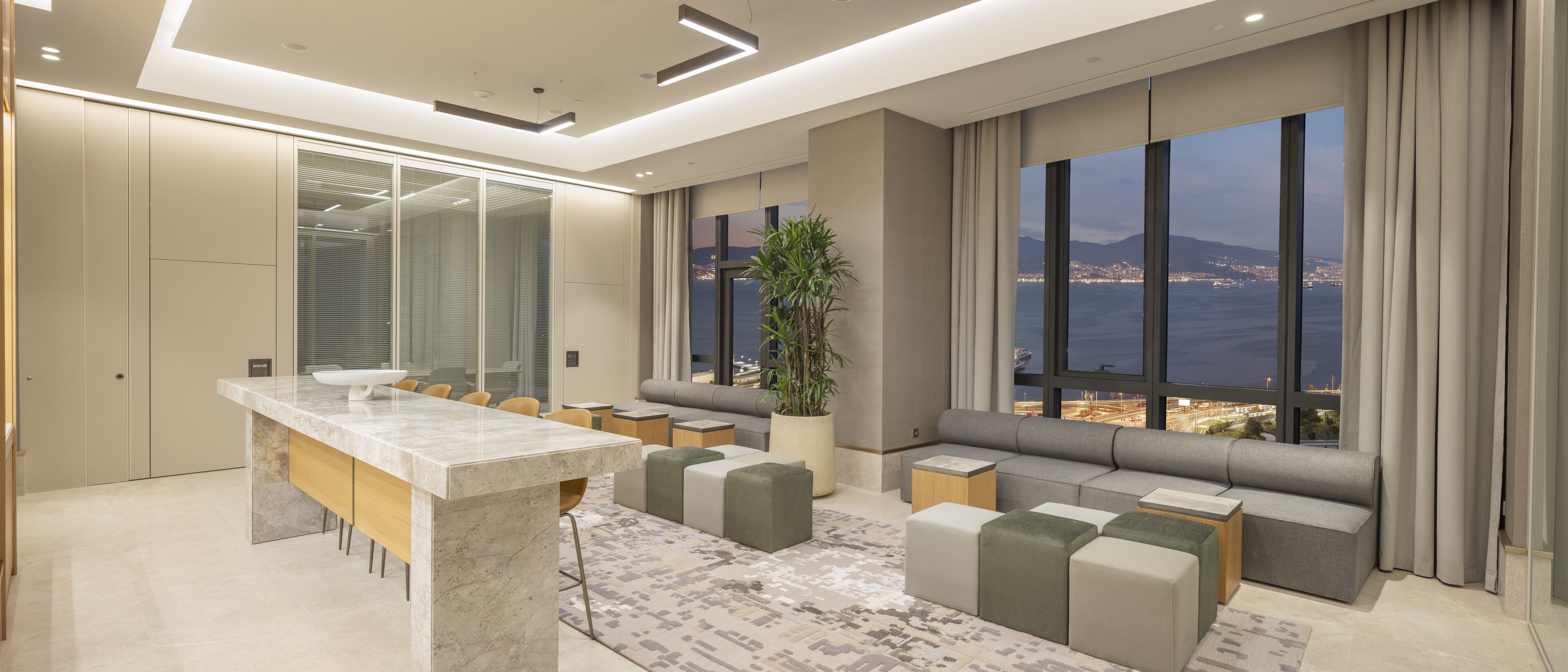 Een moderne hotelkamer met een groot raam dat uitzicht biedt op de stad, een marmeren eettafel en een zithoek met kussens.