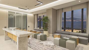 Een moderne hotelkamer met een groot raam dat uitzicht biedt op de stad, een marmeren eettafel en een zithoek met kussens.