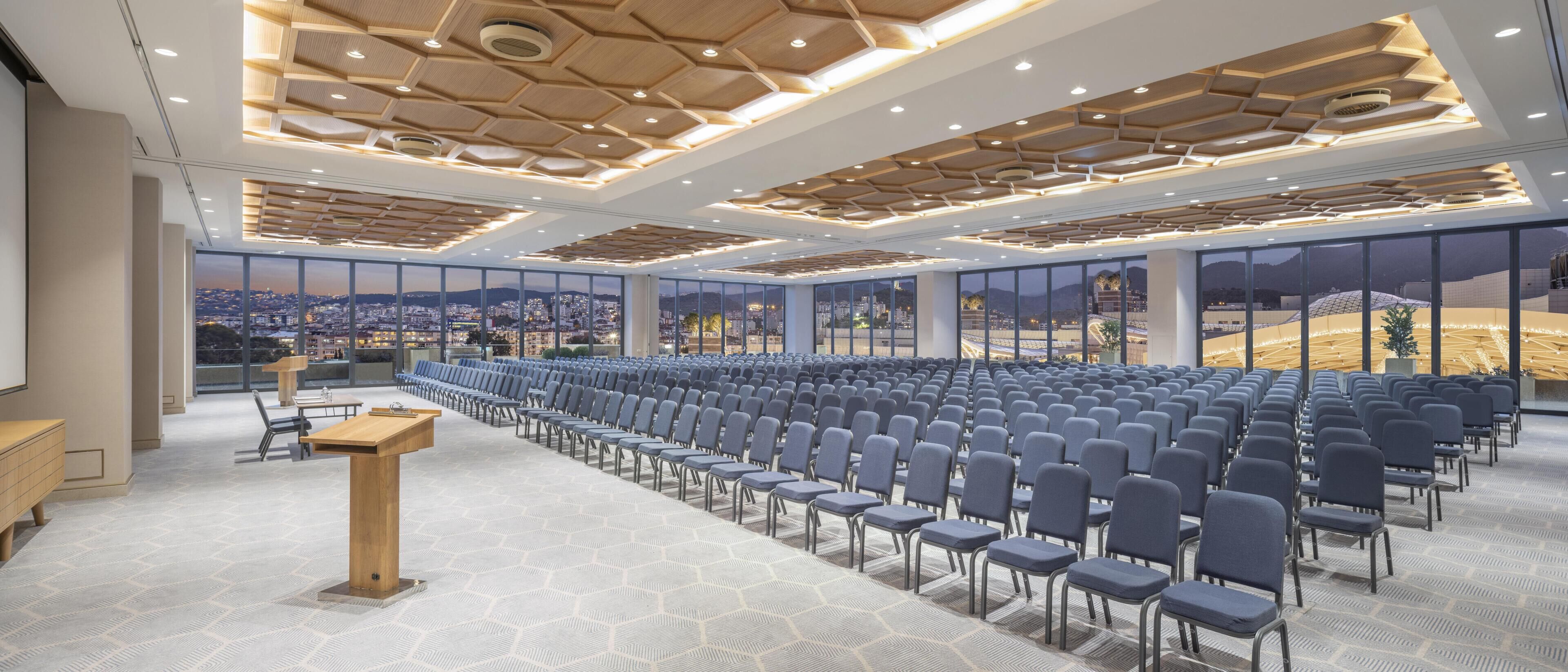 Un salón de conferencias con filas de sillas, un atril y ventanas amplias que ofrecen vistas a la ciudad.