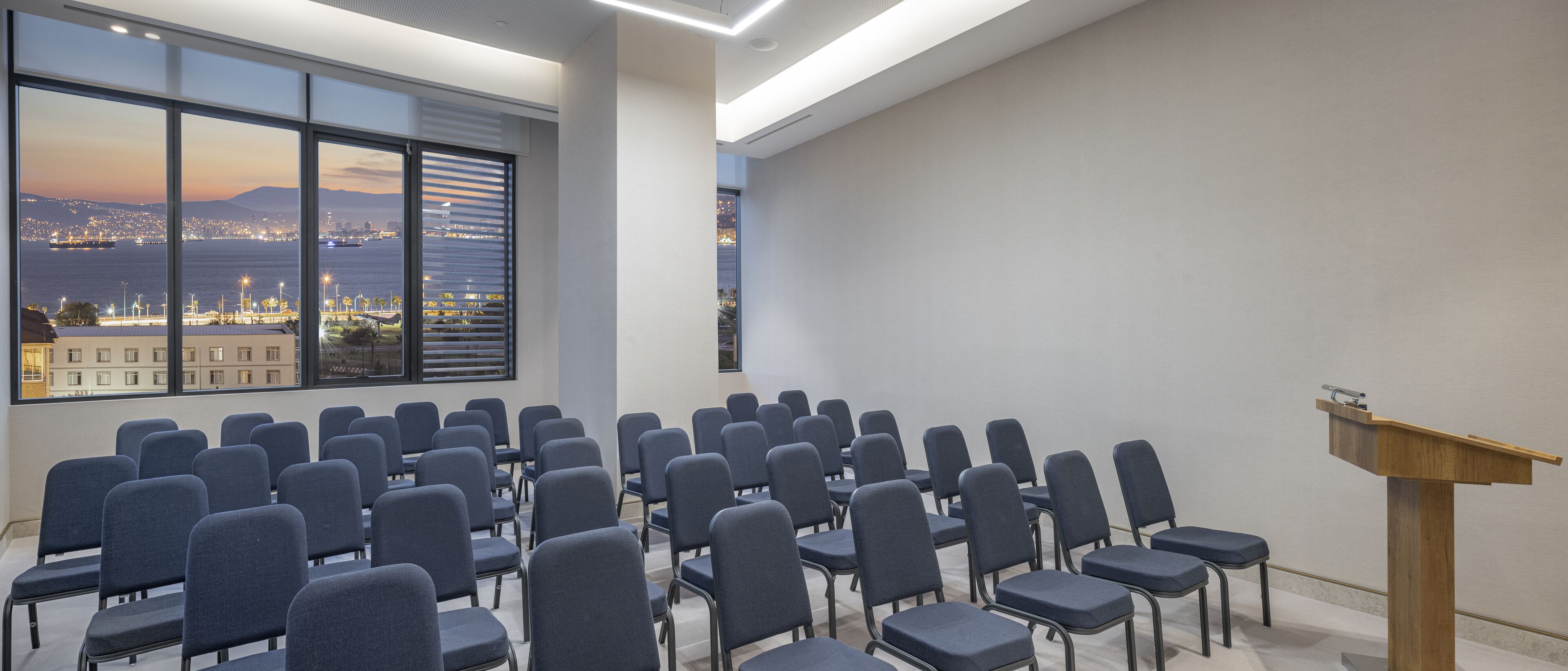 A conference room with rows of chairs and a podium, large windows with a cityscape view.