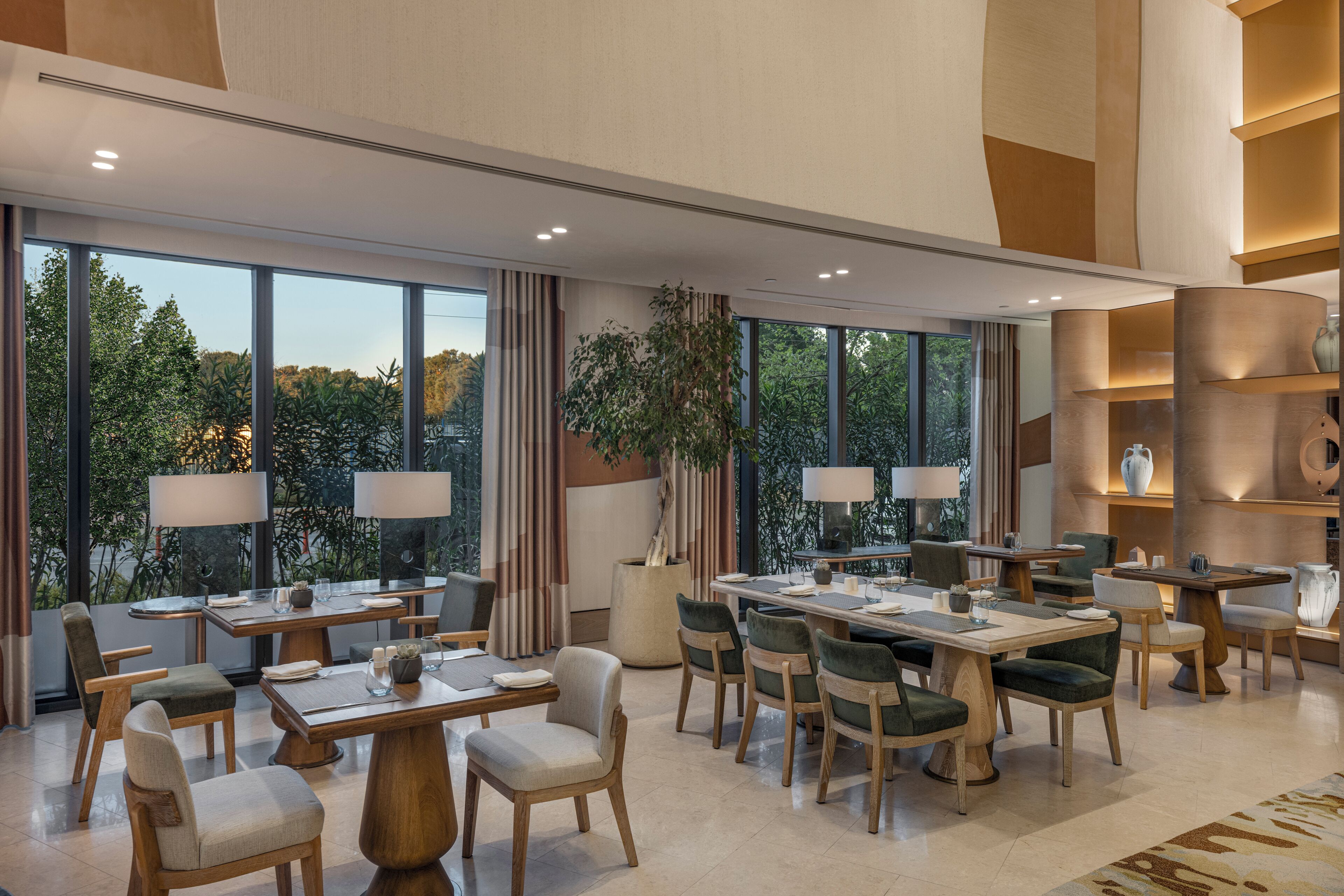 Un comedor de hotel moderno con varias mesas y sillas, ventanas amplias que dan a árboles, e iluminación ambiental.