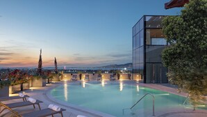 A rooftop pool with lounge chairs, a glass building, and a view of the city at dusk.