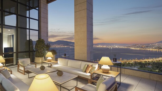 A modern outdoor lounge area with seating, tables, and lamps, overlooking a cityscape and water at dusk.