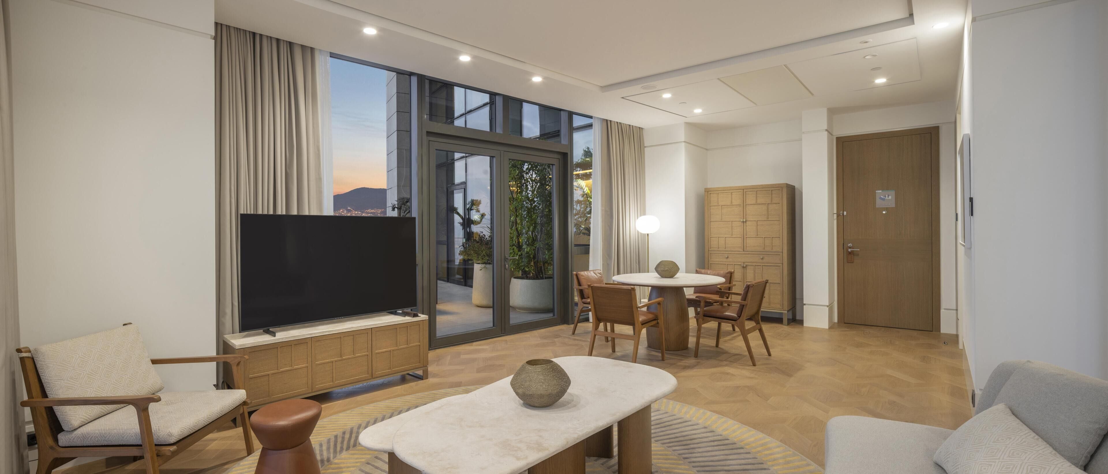 A modern living room with a flat-screen TV, a dining area, and a large window offering a city view.