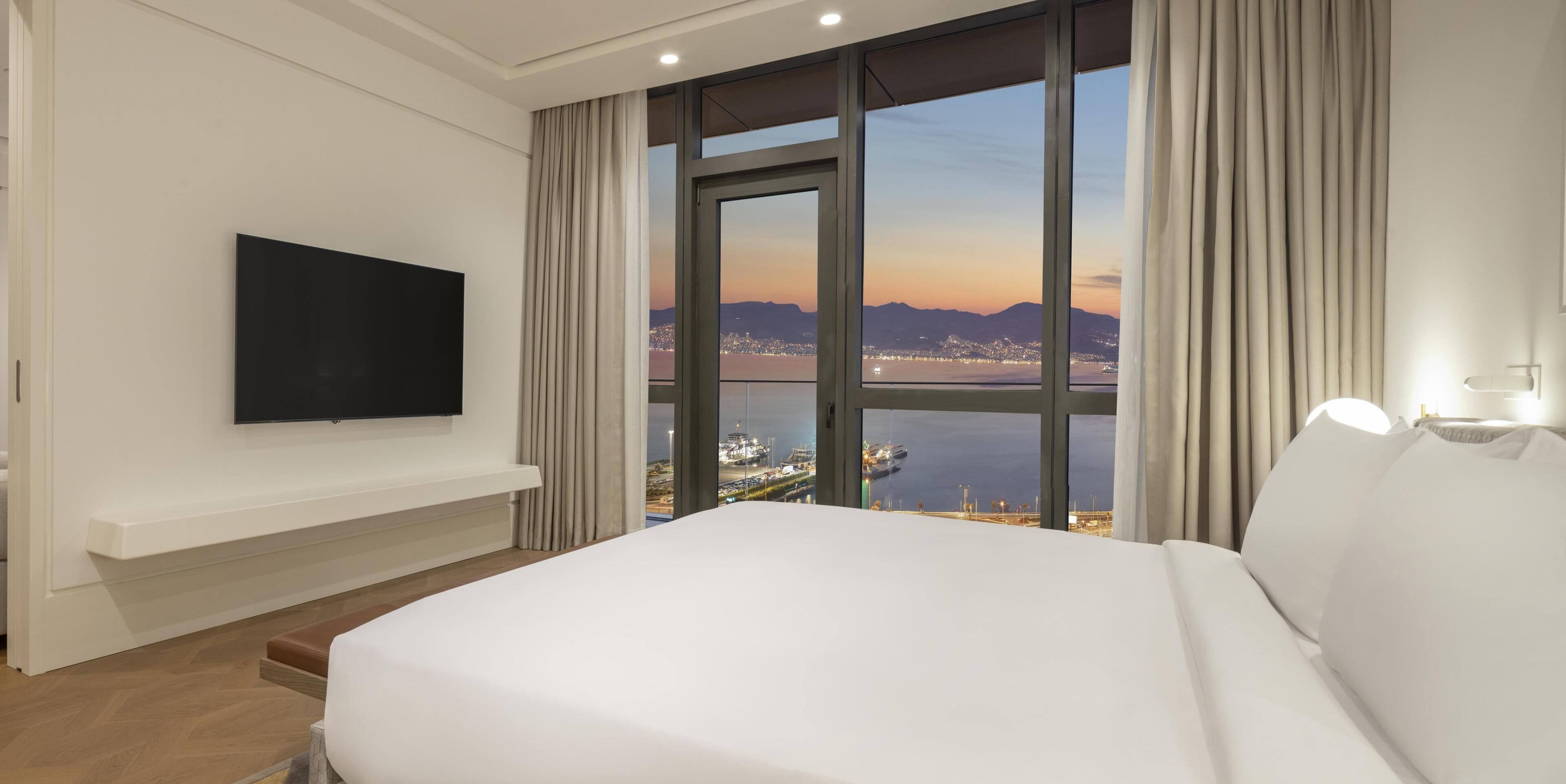 A hotel room with a large bed, a flat-screen TV mounted on the wall, and a view of a cityscape and body of water through large windows.