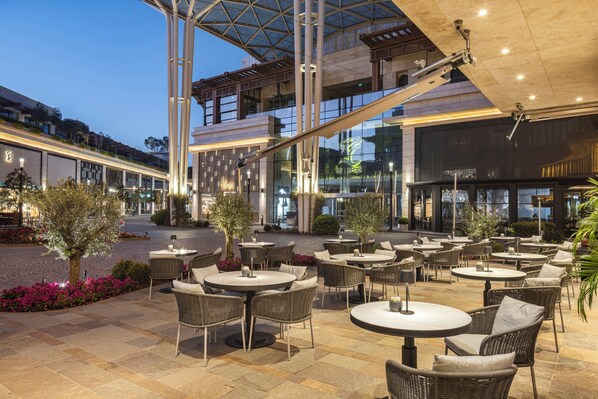 An outdoor patio area with round tables and wicker chairs, surrounded by potted plants and a modern architectural structure. - Grand Hyatt Izmir Istinyepark (Izmir)