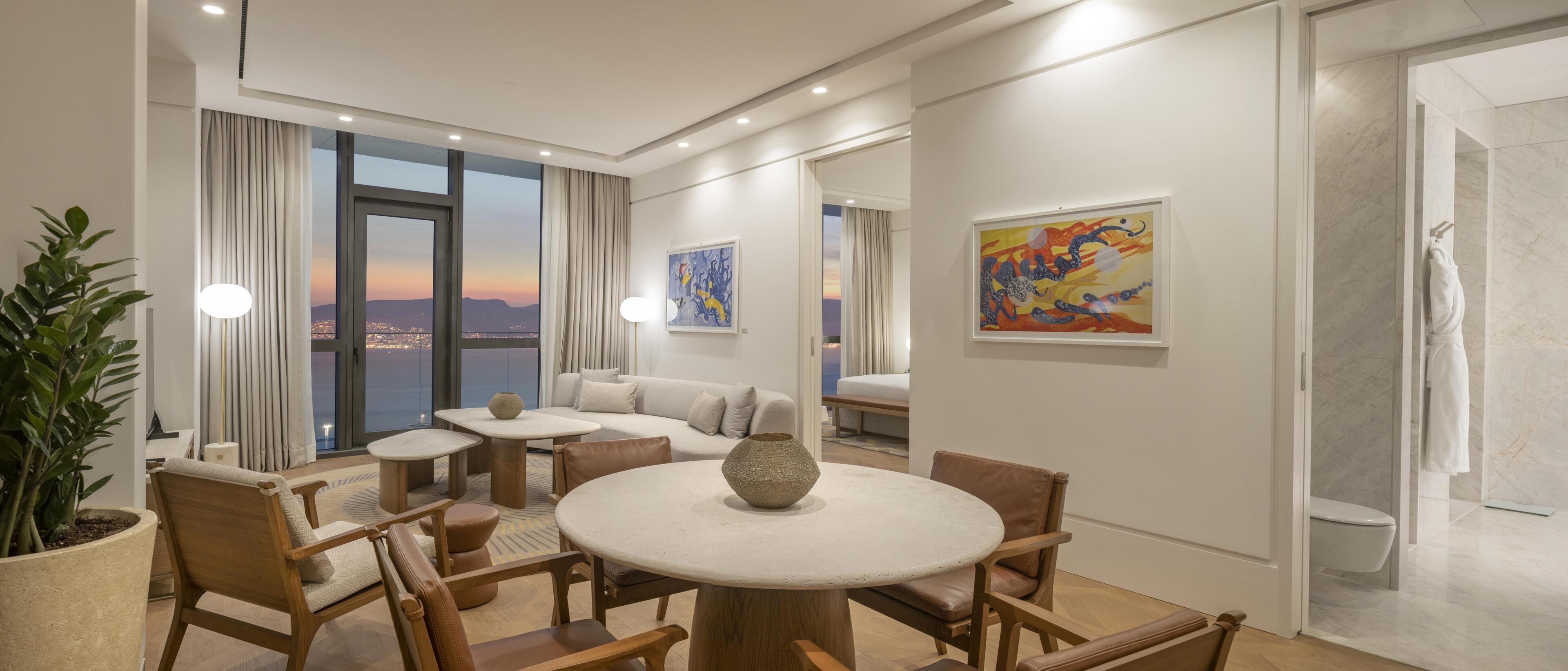 Una habitación de hotel moderna con comedor, sofá, ventana grande con vistas a la ciudad y obras de arte en las paredes.
