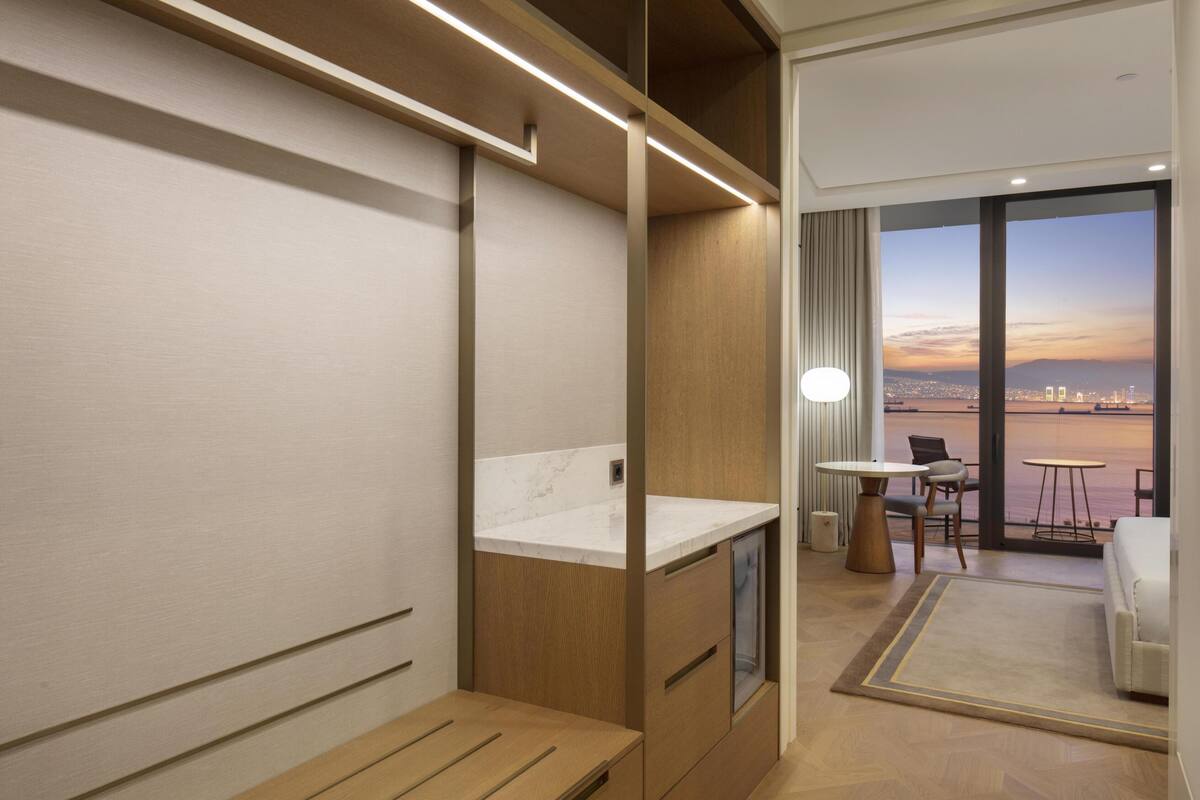 Een moderne hotelkamer met een geïntegreerde keuken, een balkon met uitzicht op de stad, en een zithoek met een tafel en stoelen.