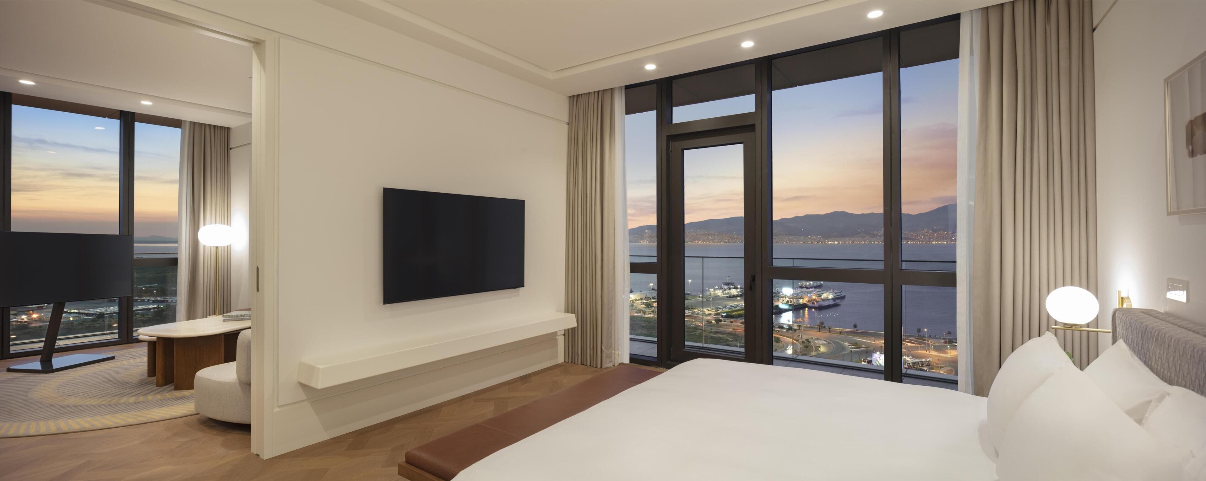 A hotel room with a large bed, a flat-screen TV, a desk, and a view of a cityscape through large windows.