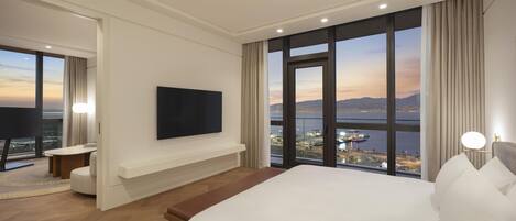 A hotel room with a large bed, a flat-screen TV, a desk, and a view of a cityscape through large windows.