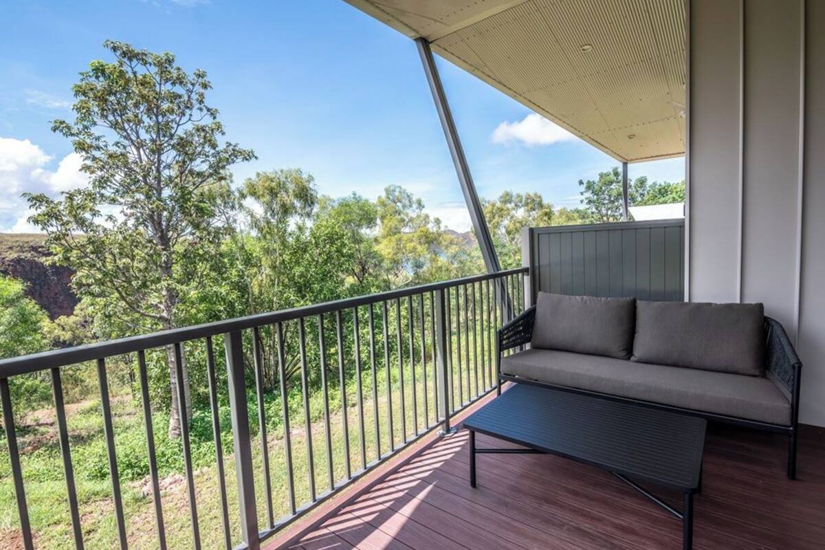 Ein Balkon mit einer Couch und einem Tisch, mit Blick auf eine natürliche Landschaft mit Bäumen und blauem Himmel.