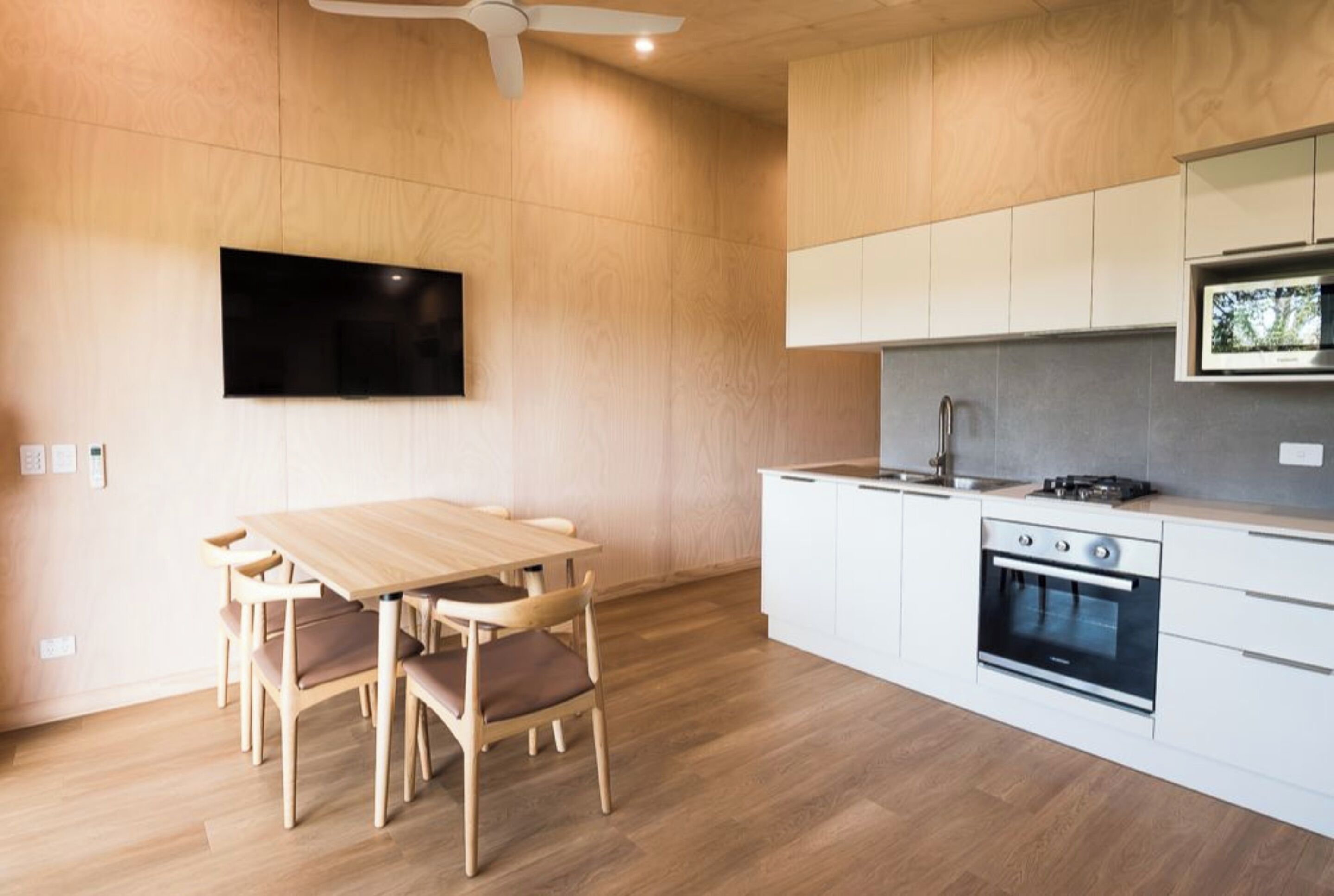 A modern kitchen with a dining table and chairs, a built-in oven, and a flat-screen TV mounted on the wall.
