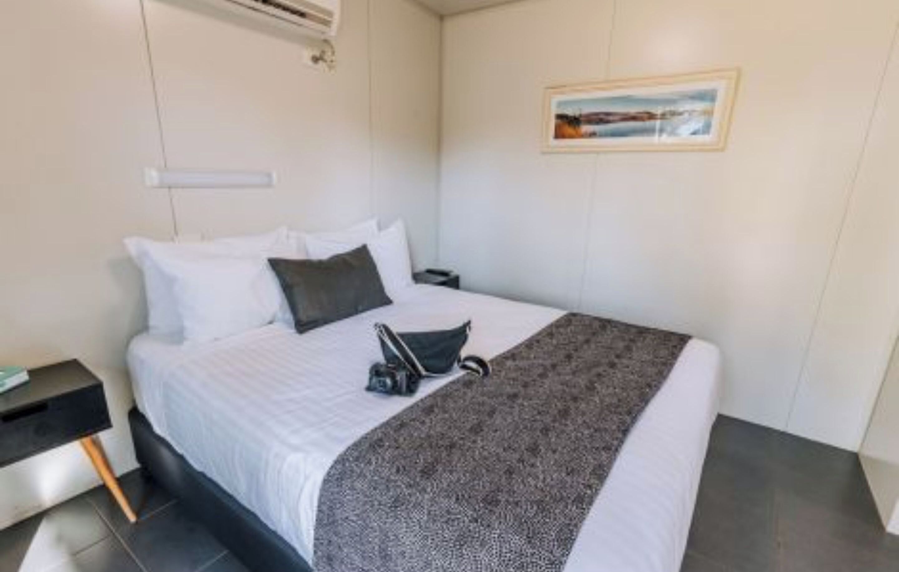 A neatly made bed with a white comforter and a patterned throw blanket, a black nightstand with a lamp, and a framed picture on the wall.