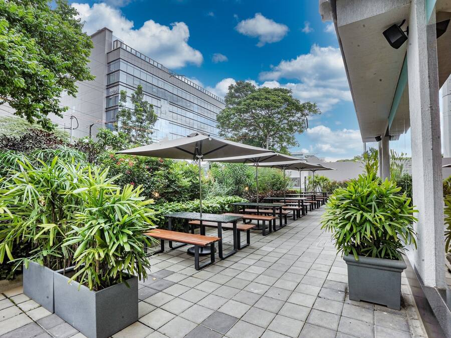 An outdoor patio area with seating and planters, under a covered structure, adjacent to a modern building.