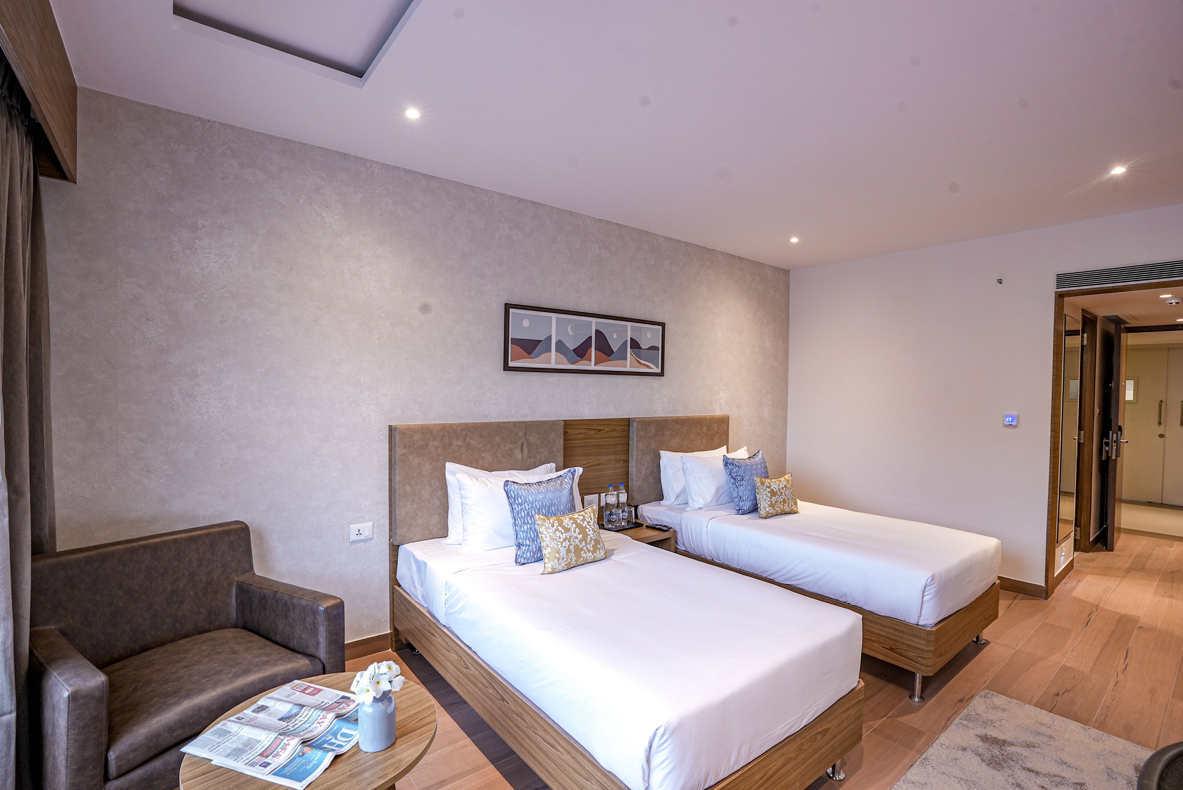 A hotel room with two beds, a sofa, a small table with a newspaper, and a painting on the wall.