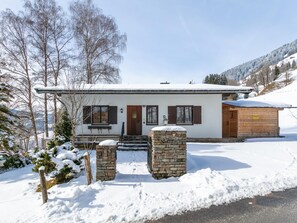 A two-story chalet with a stone entrance, wooden shutters, and a snow-covered roof.