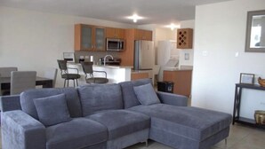 Ein modernes Wohnzimmer mit einem grauen Ecksofa, einem Essbereich mit Tisch und Stühlen sowie einer Küche mit Holzschränken und Geräten.