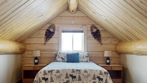 A cozy attic bedroom with a sloped ceiling, a large window, and a bed with a moose-themed duvet. - Beautiful hand-crafted log cabin on Horse Creek one block off Main Street. (Dubois)