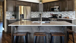 A modern kitchen with dark wood cabinets, stainless steel appliances, a large island with bar stools, and a wooden ceiling. - Beautiful hand-crafted log cabin on Horse Creek one block off Main Street. (Dubois)