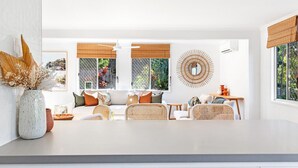 Ein modernes Wohnzimmer mit einem weißen Sofa, Korbstühlen und einem Esstisch. Große Fenster mit Bambusrollos, ein dekorativer Spiegel und eine Vase mit Trockenblumen sind ebenfalls zu sehen.