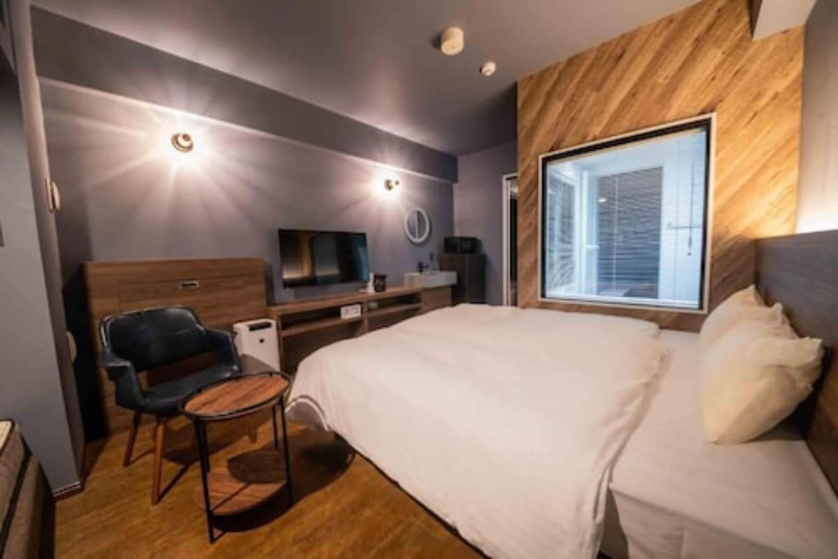 A modern hotel room with a large bed, a desk with a chair, a television, and a large window with blinds.