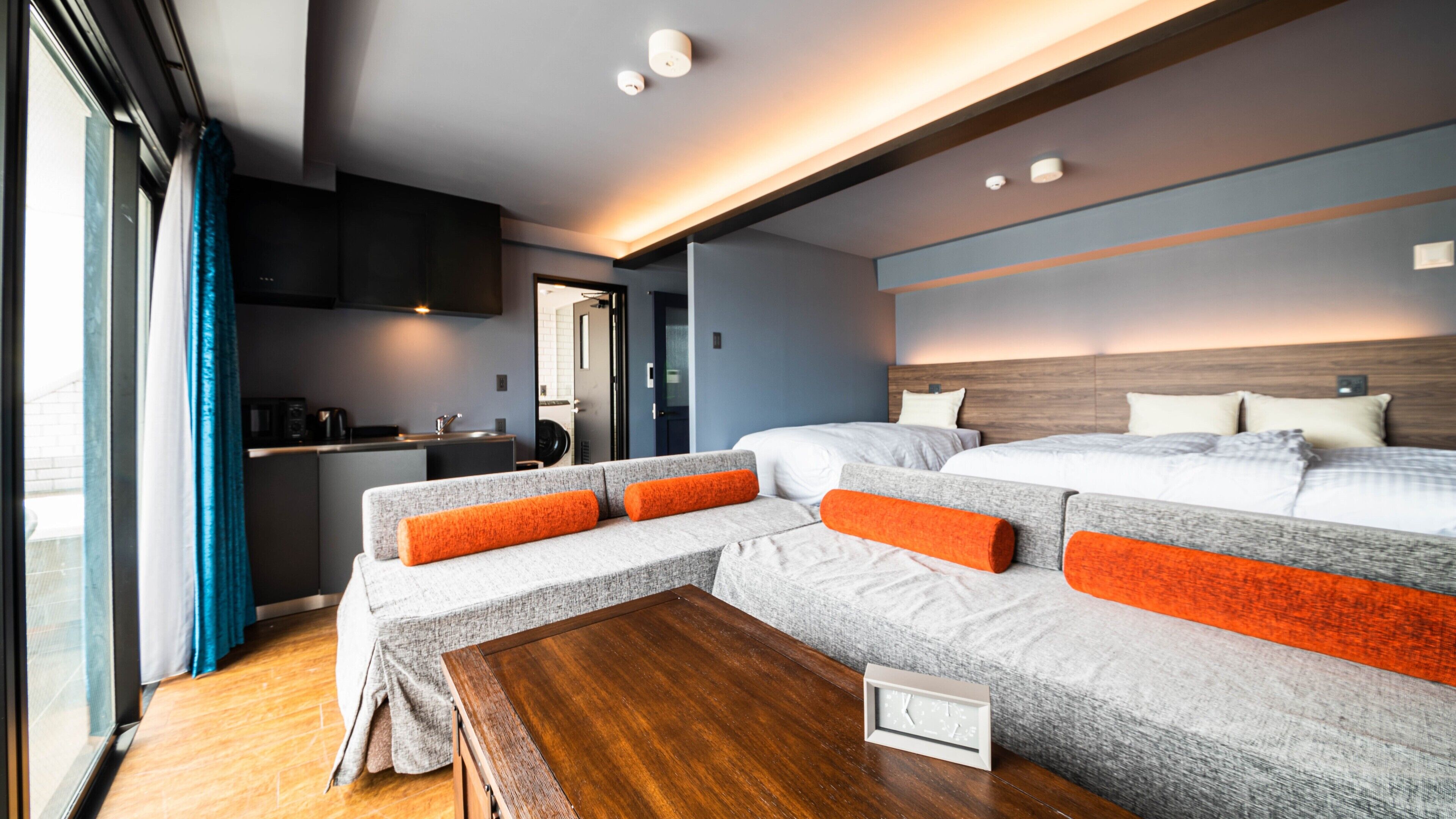 A modern hotel room with a sofa bed, a wooden table, a kitchen area, and a large window.
