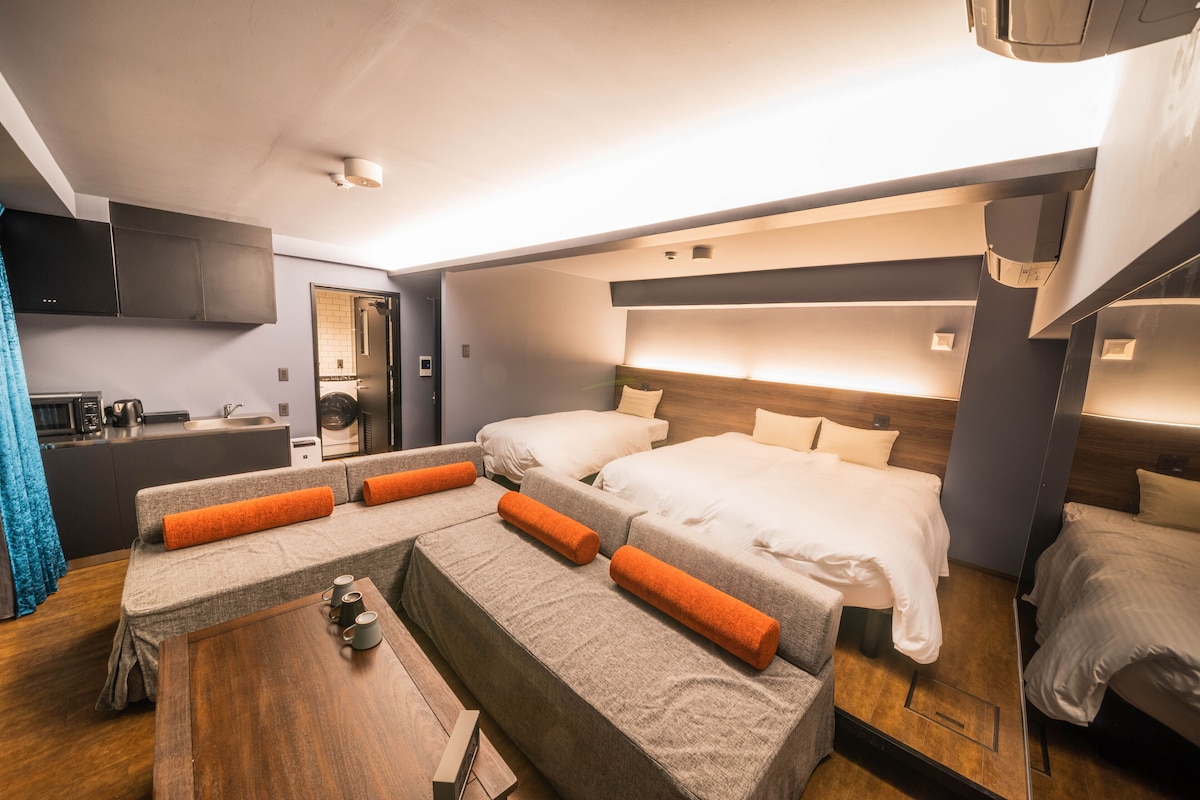 A modern hotel room with two beds, a sofa, a wooden table, and a kitchenette.