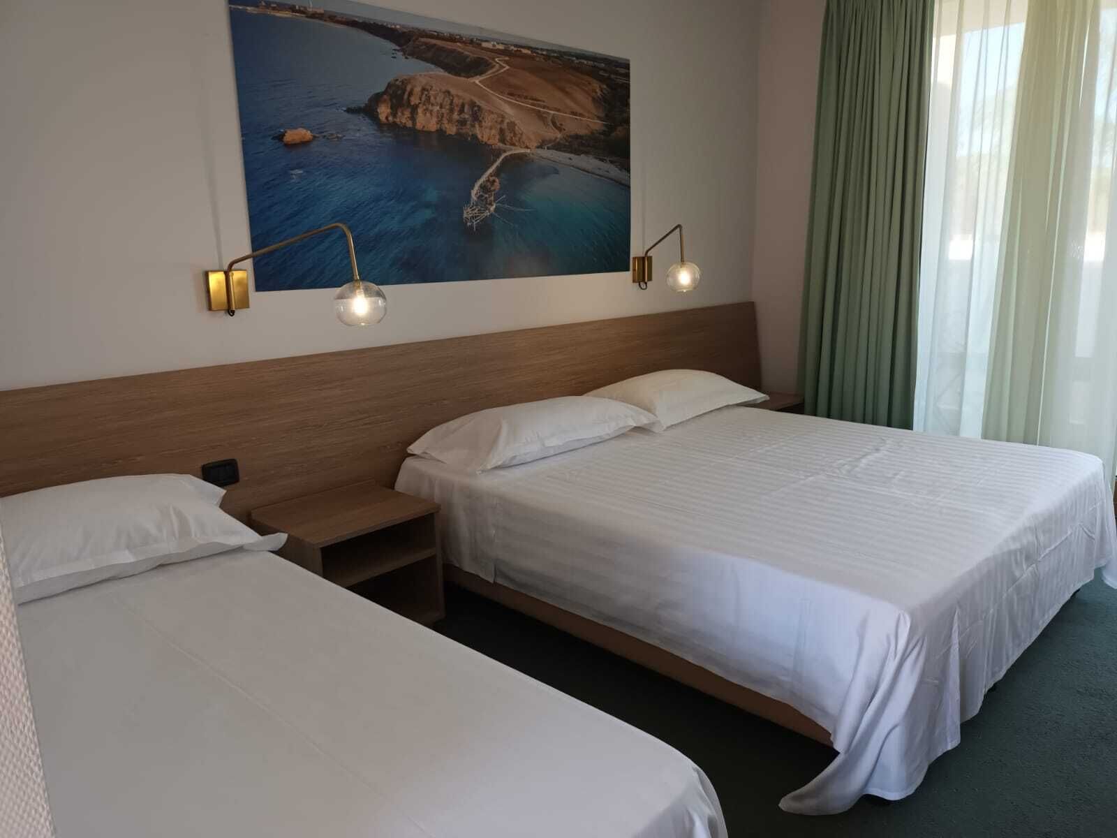 A hotel room with two single beds, a wooden headboard, a nightstand, a wall-mounted picture of a coastal landscape, and a window with curtains.