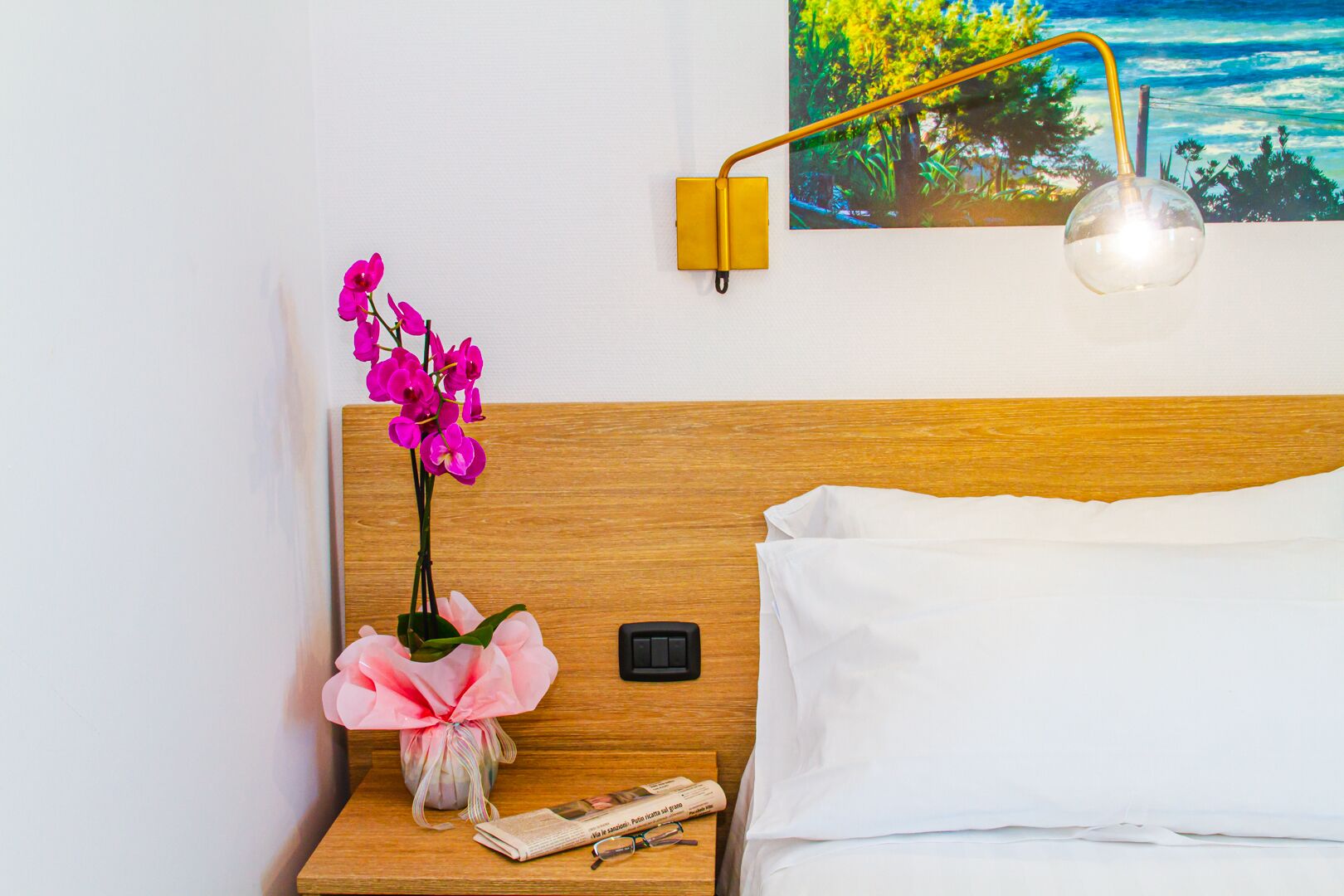 Cama bem arrumada com lençóis brancos, cabeceira de madeira, mesa de cabeceira com abajur, um vaso com flores cor-de-rosa e uma pintura de uma paisagem.
