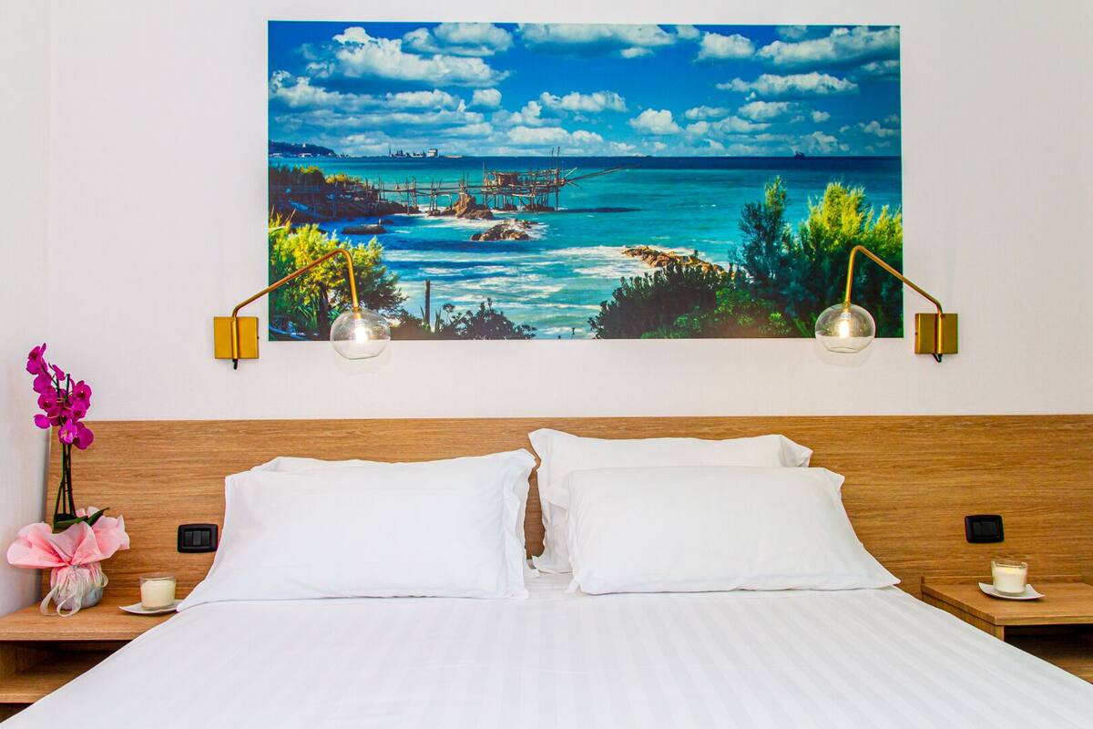 A hotel room with a large bed, two bedside tables, a nightstand, a wall-mounted painting of a coastal scene, and two wall-mounted lamps.