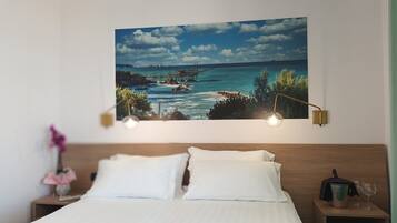 A bedroom with a large ocean view painting, a wooden headboard, white bedding, a bedside table with a lamp, and a small nightstand with a vase of flowers.