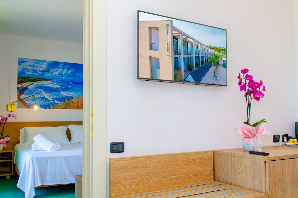 A hotel room with a bed, a desk, a chair, a small table with a vase of flowers, and a painting of a building on the wall.