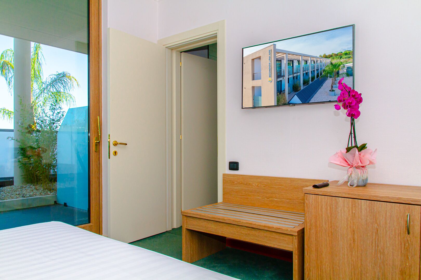 A hotel room with a bed, a wooden bench, a desk, a mirror, and a window with a view of a building and greenery.