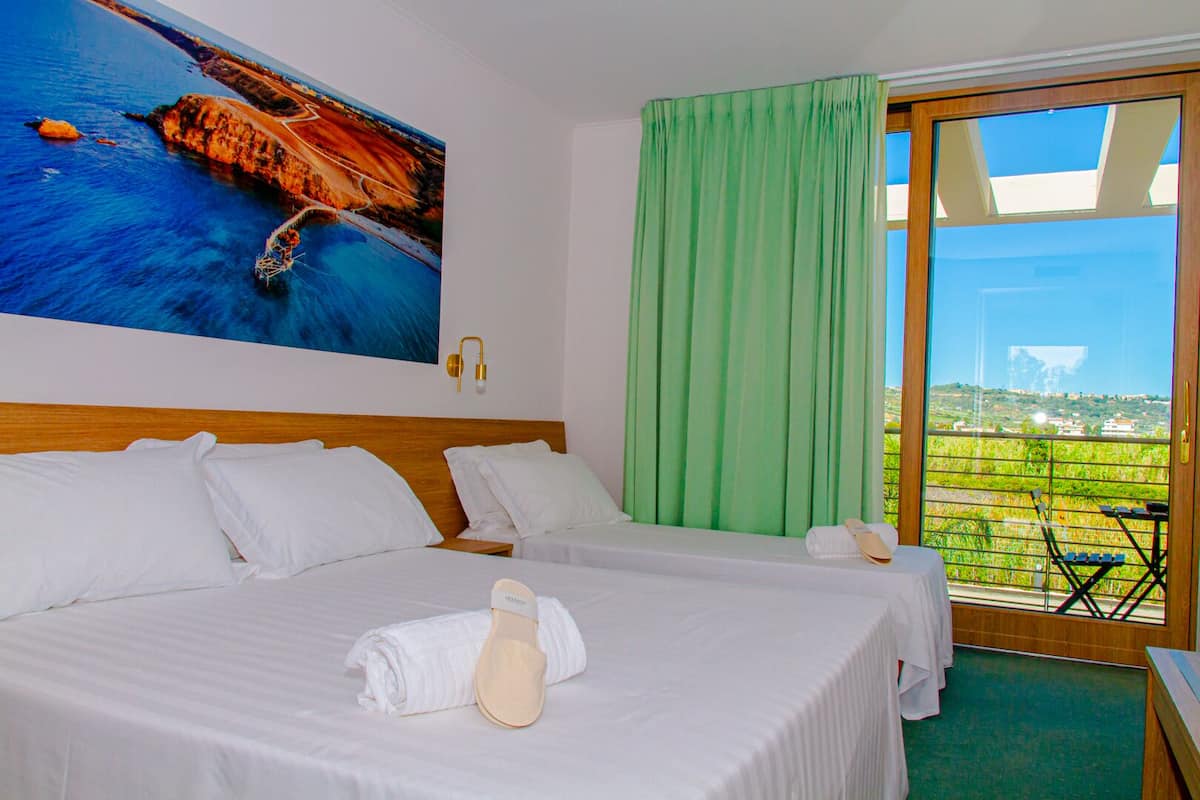 A hotel room with two beds, a large painting of a coastal landscape, a green curtain, and a view of a green field through a glass door.
