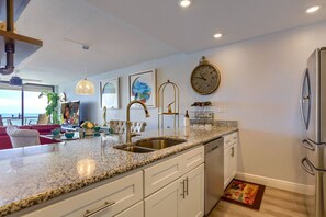 A modern kitchen with a granite countertop, white cabinetry, a stainless steel sink, and a built-in oven. There are framed artworks on the wall and a large clock above the sink.