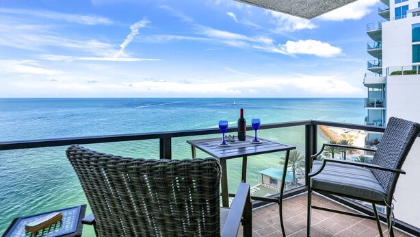 Een balkon met een tafel, twee stoelen en een fles wijn. In de verte is de oceaan en een kustlijn te zien.