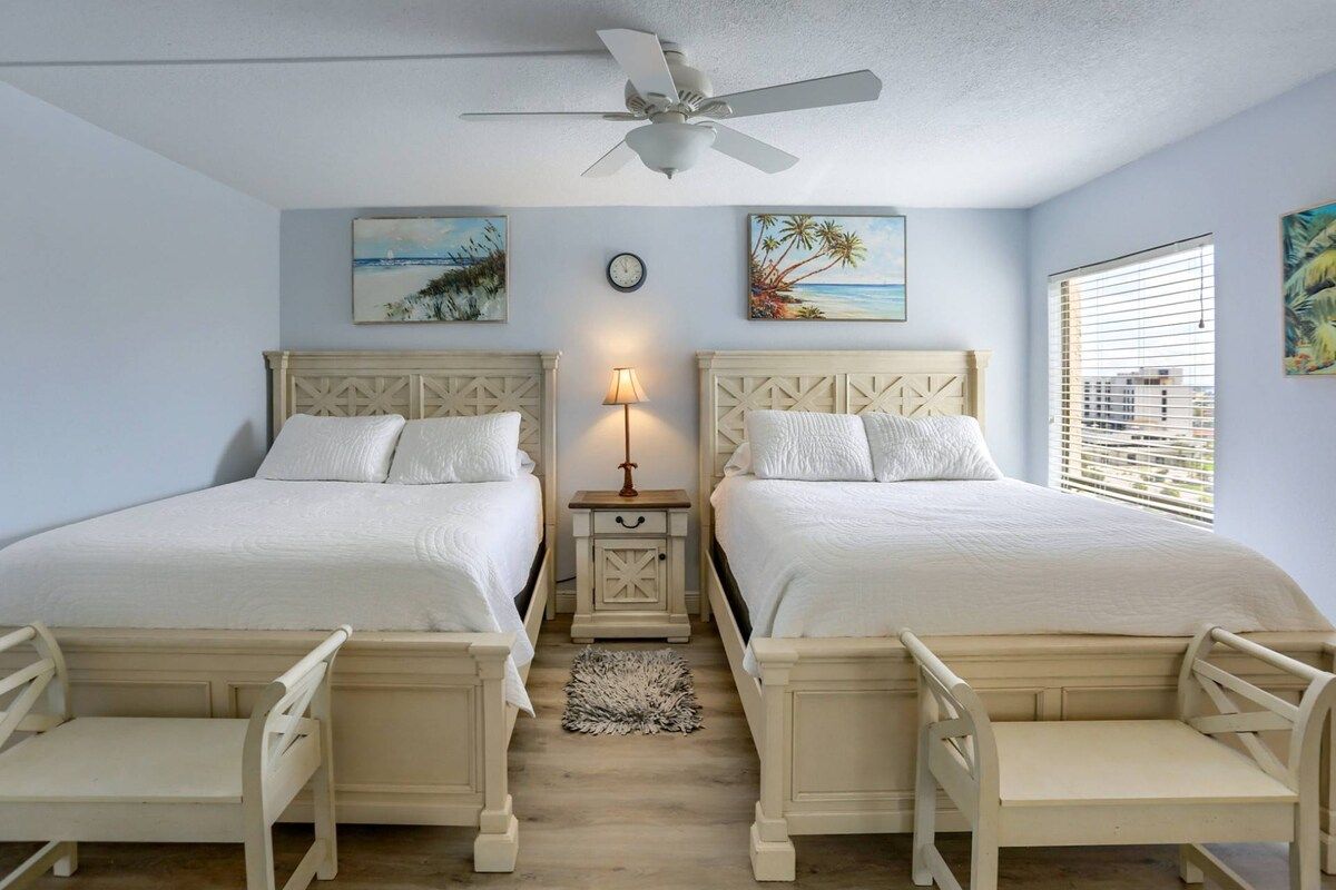 A room with two beds, a ceiling fan, artwork on the walls, a clock, and a window with blinds.